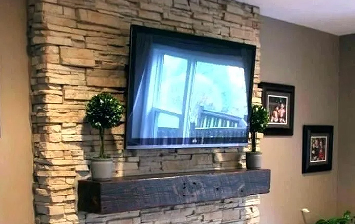 Flat-screen TV mounted on a stone accent wall with a wooden mantel and two potted plants.
